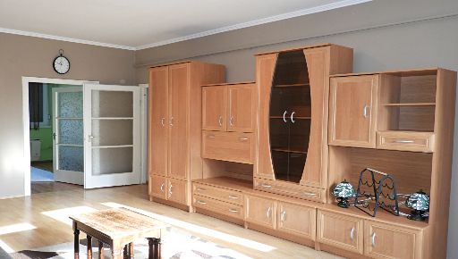 Ezen a fotón egy nappali szoba belsőt látunk, sarkosan fotózva, a kávébarna falak előtt fiatalos szekrénysorral, fehér mennyezettel. Látjuk a másik szobába vezető, nyitott ajtót is.