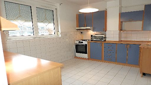 Ezen a fotón egy tágas konyhát látunk, sarkosan fotózva, bal oldalon két, redőnyös ablakkal, rájuk merőlegesen konyhaszekrénnyel, tűzhellyel, alsó-felső elemekkel, munkapulttal.